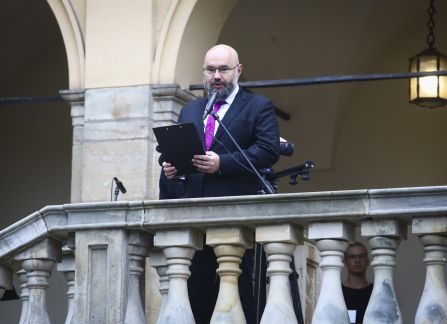 Fotografia. Mężczyzna w garniturze przemawia ze szczytu schodów, stojąc za kamienną balustradą.
