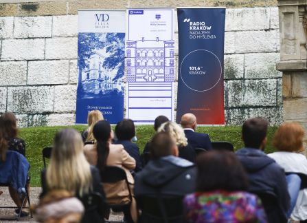 Fotografia. Publiczność wydarzenia plenerowego. Ludzie siedzą na rozkładanych krzesłach. W tle fasada willi Decjusza.