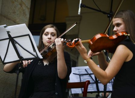 Fotografia. Dwie kobiety występują. Grają na skrzypcach. W tle fragment fasady Willi Decjusza.