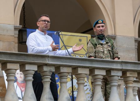 Fotografia. Mężczyzna w białej koszuli przemawia ze szczytu schodów przed Willą Decjusza. Obok stoi mężczyzna w mundurze wojskowym.