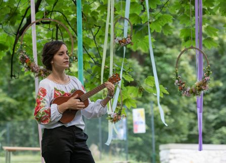 Fotografia. Kobieta w haftowanej na kolorowo bluzce gra na ukulele. W tle drzewa.