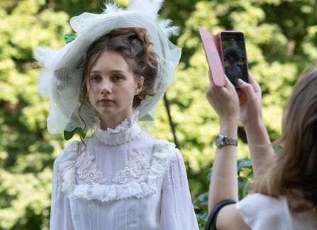 Fotografia. Kobieta w rustykalnej białej sukni i białym kapeluszu pozuje do zdjęcia na tle drzew.