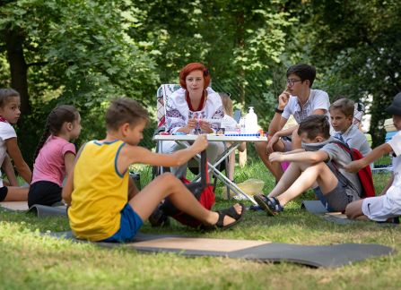 Fotografia. Kobieta w zdobionej, ludowej bluzce prowadzi zajęcia plastyczne na zewnątrz. Dzieci siedzą na trawie.