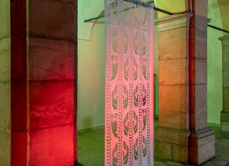 a photograph. large-format cutout with a traditional pattern, displayed between the columns of arcades on the facade of the Decius villa. Photo taken after dark, the cut-out highlighted in green and red.