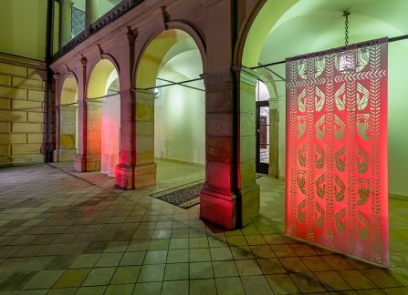 a photograph. large-format cutout with a traditional pattern, displayed between the columns of arcades on the facade of the Decius villa. Photo taken after dark, the cut-out highlighted in green and red.